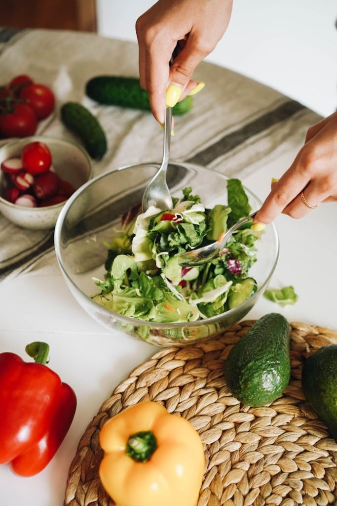 Hands preparing a fresh vegetable salad with cucumbers, peppers, and lettuce in a kitchen setting.